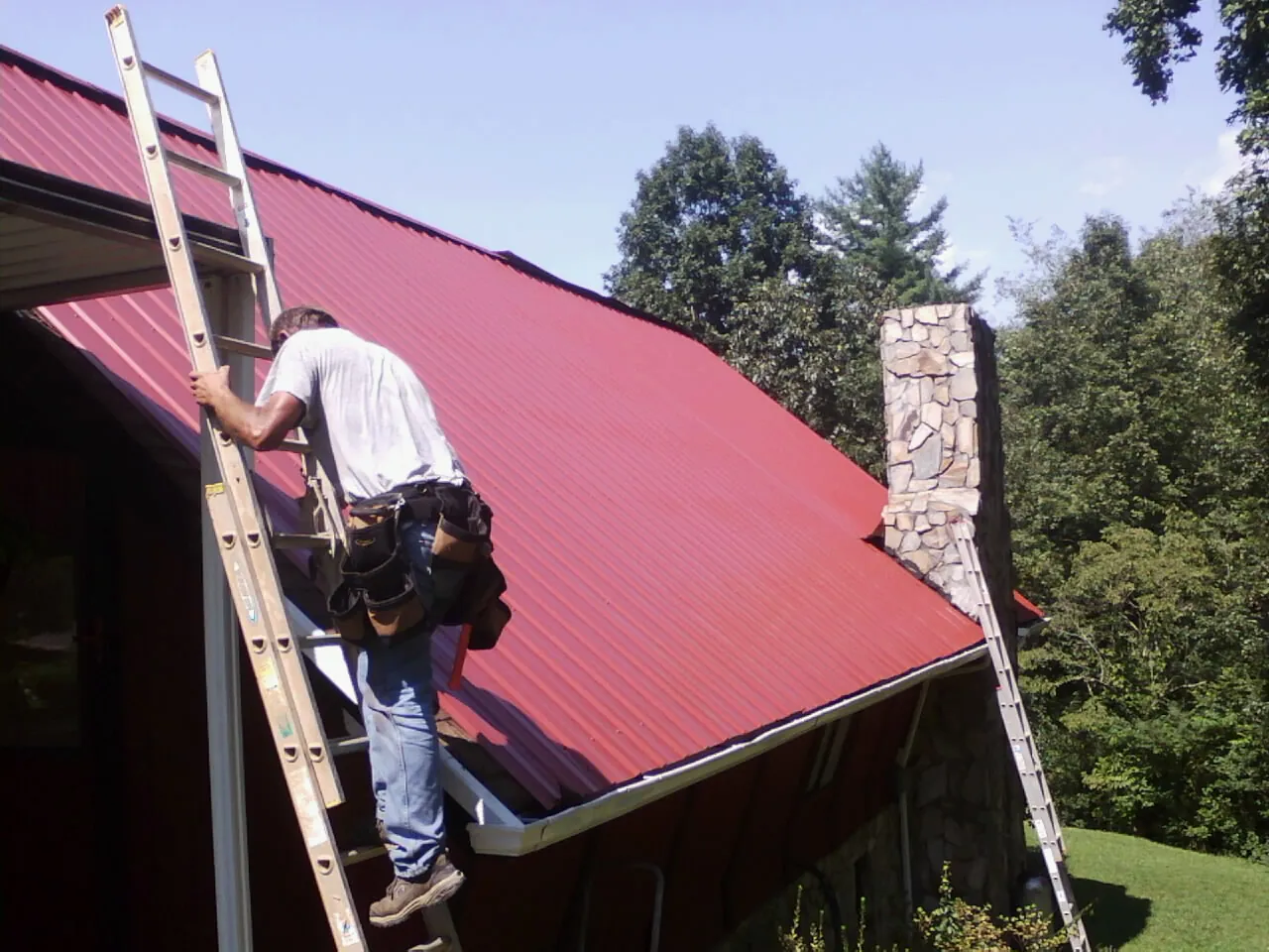 Roofer on a ladder beside a red metal roof and stone chimney — Soffit & Fascia Repair by Roofing Brothers in Randle
